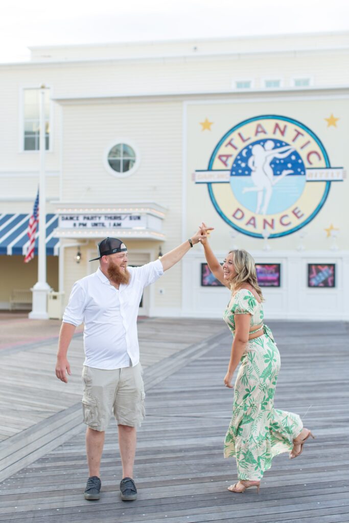 Guy twirls laughing Girl as they dance in front of the Dance Hall at the Boardwalk for their Disney Engagement Photos