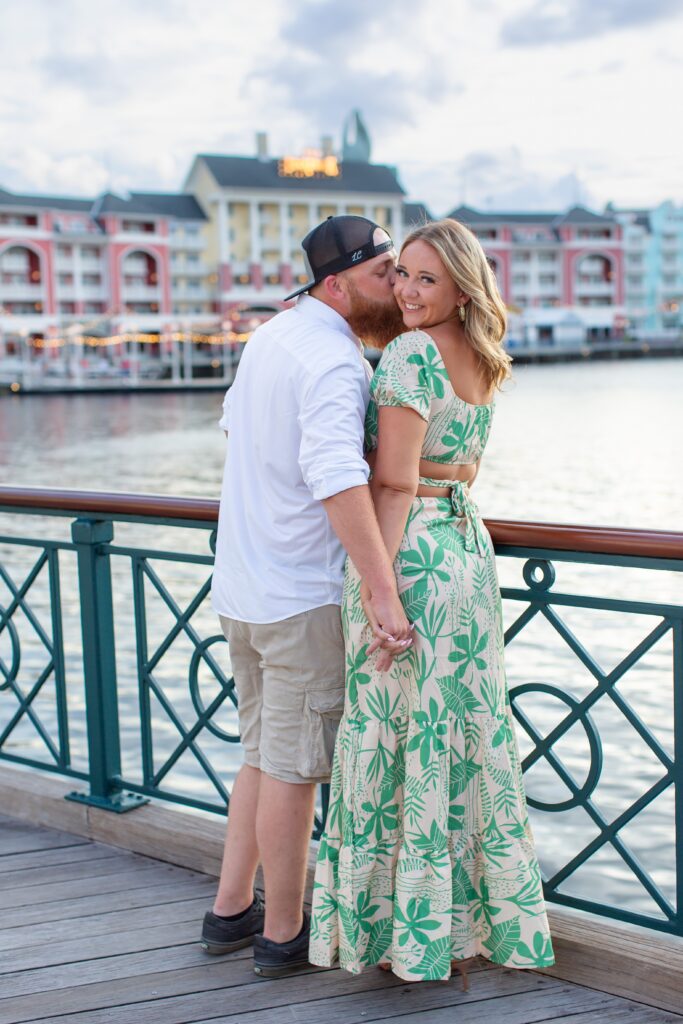 Couple holds hands and guy kisses her cheek as they stand in front of the water at the Boardwalk for their Disney Engagement Photos