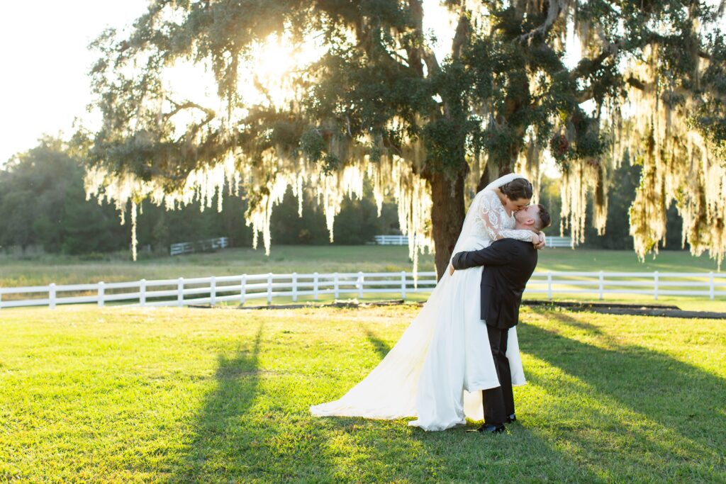 Groom lifts Bride under large tree at sunset in open field at Bramble Tree Estate in Orlando, Florida