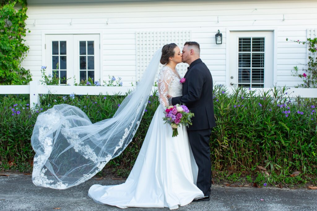 Bride and Groom kiss in front of flowers and white building while Bride's veil flys in the wind at Bramble Tree Estate in Orlando, Florida