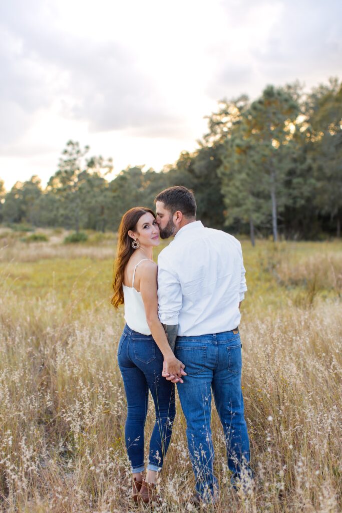 Couple holding hands while guy kisses girl's cheek standing in field for their engagement photos at Lake Louisa State Park in Orlando Florida