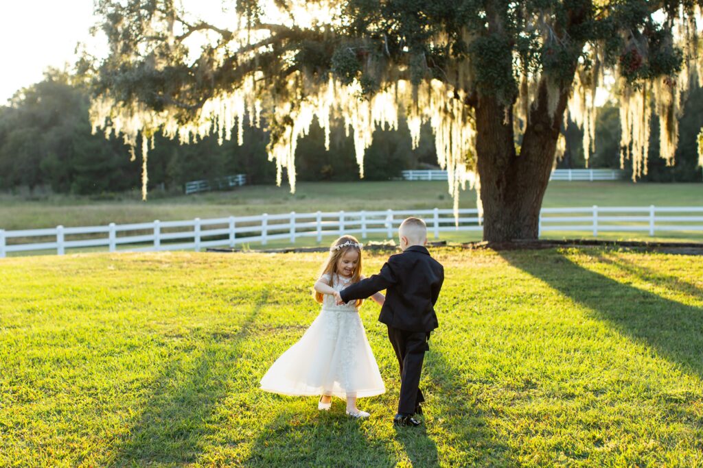 Flower girl and ring bearer hold hands and spin under large tree at sunset in open field at Bramble Tree Estate in Orlando, Florida