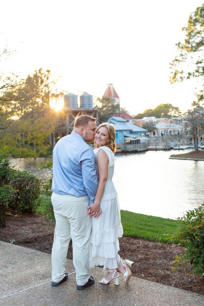 Couple holds hands as guy kisses her cheek in front of Riverside scene at sunset for their Disney Engagement Photos at Port Orleans Resort