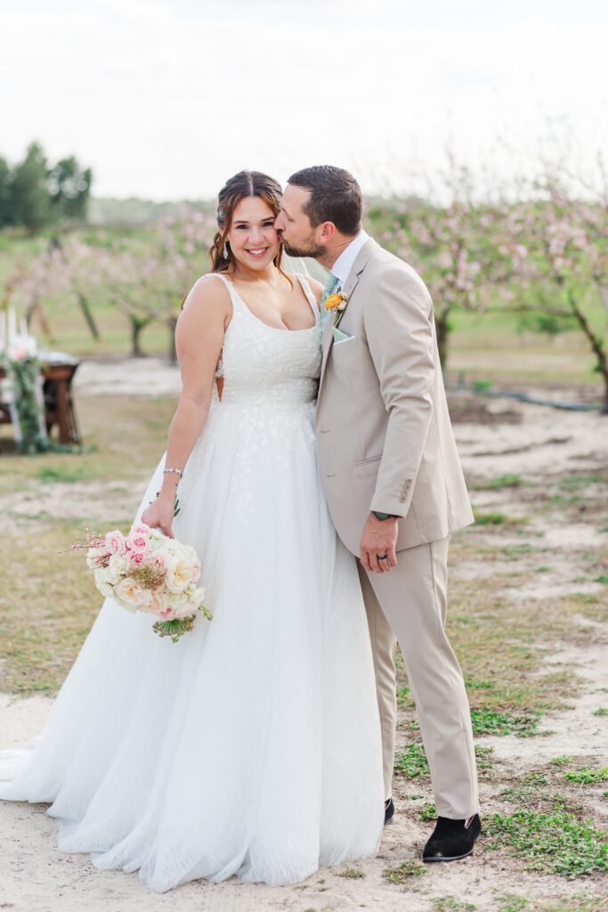 Groom kisses Bride's cheek standing in the peach blossoms after their wedding at Acres of Grace Wedding Venue in Central Florida
