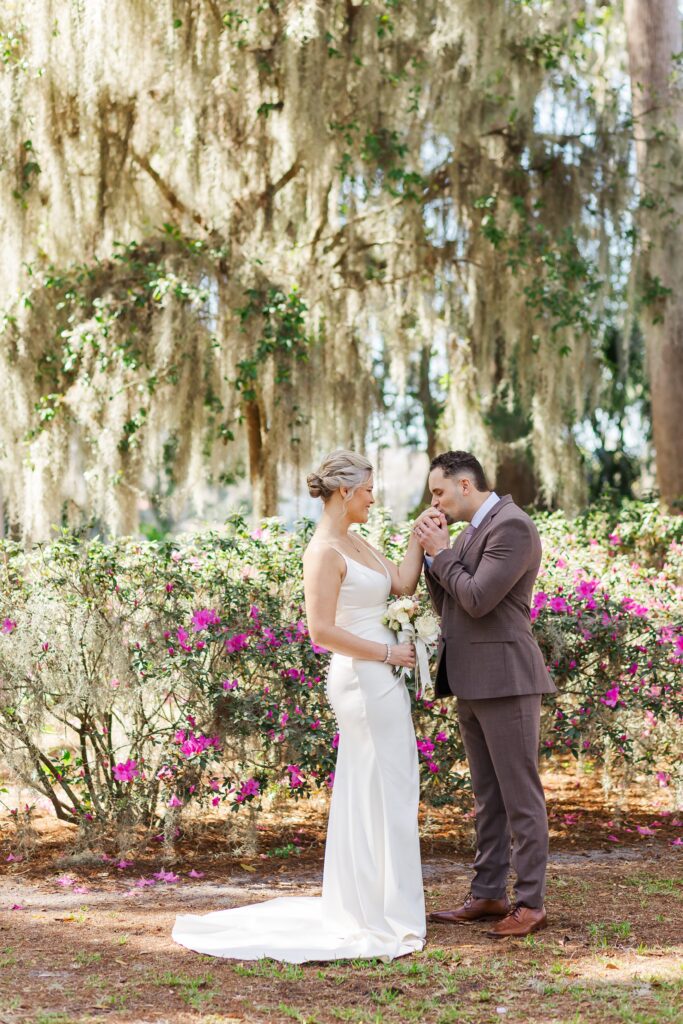 Groom kisses Bride's hand by purple azaleas under beautiful live oak trees and moss at Kraft Azalea Garden Wedding in Orlando, Florida