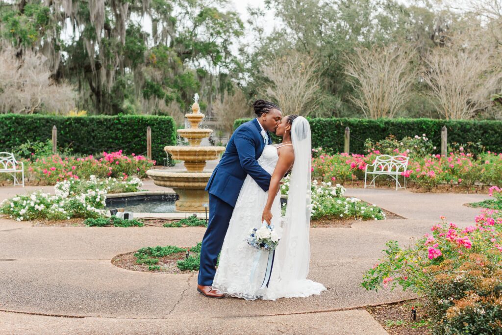 Groom dips and kisses bride in blooming rose garden with fountain behind them after their elopement at Leu Gardens in Orlando, Florida