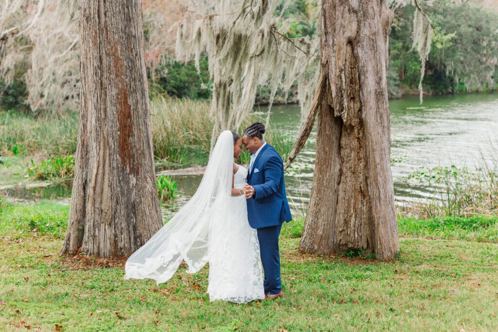Bride and Groom hold hands standing in front of lake and like oak trees with moss after their elopement at Leu Gardens in Orlando, Florida