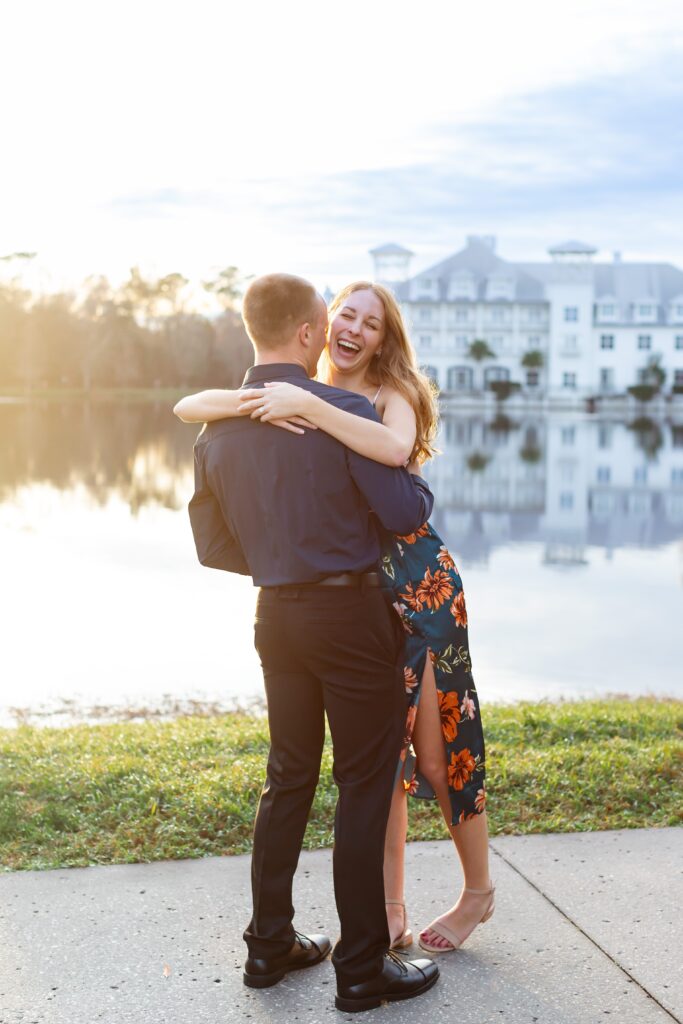 Girl wraps arms around guy and laughs as he swings her by the water in front of the Celebration Bohemian Hotel for their engagement photos at sunset in Celebration, Florida
