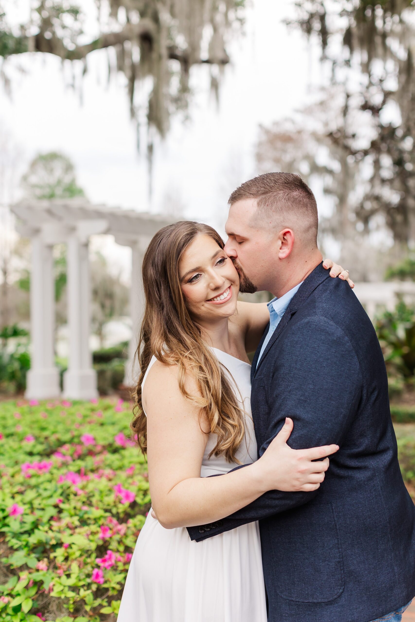 Guys kisses girl's cheek in front of beautiful spanish moss and flowers for their Orlando Engagement Photos at Cypress Grove Park in Orlando