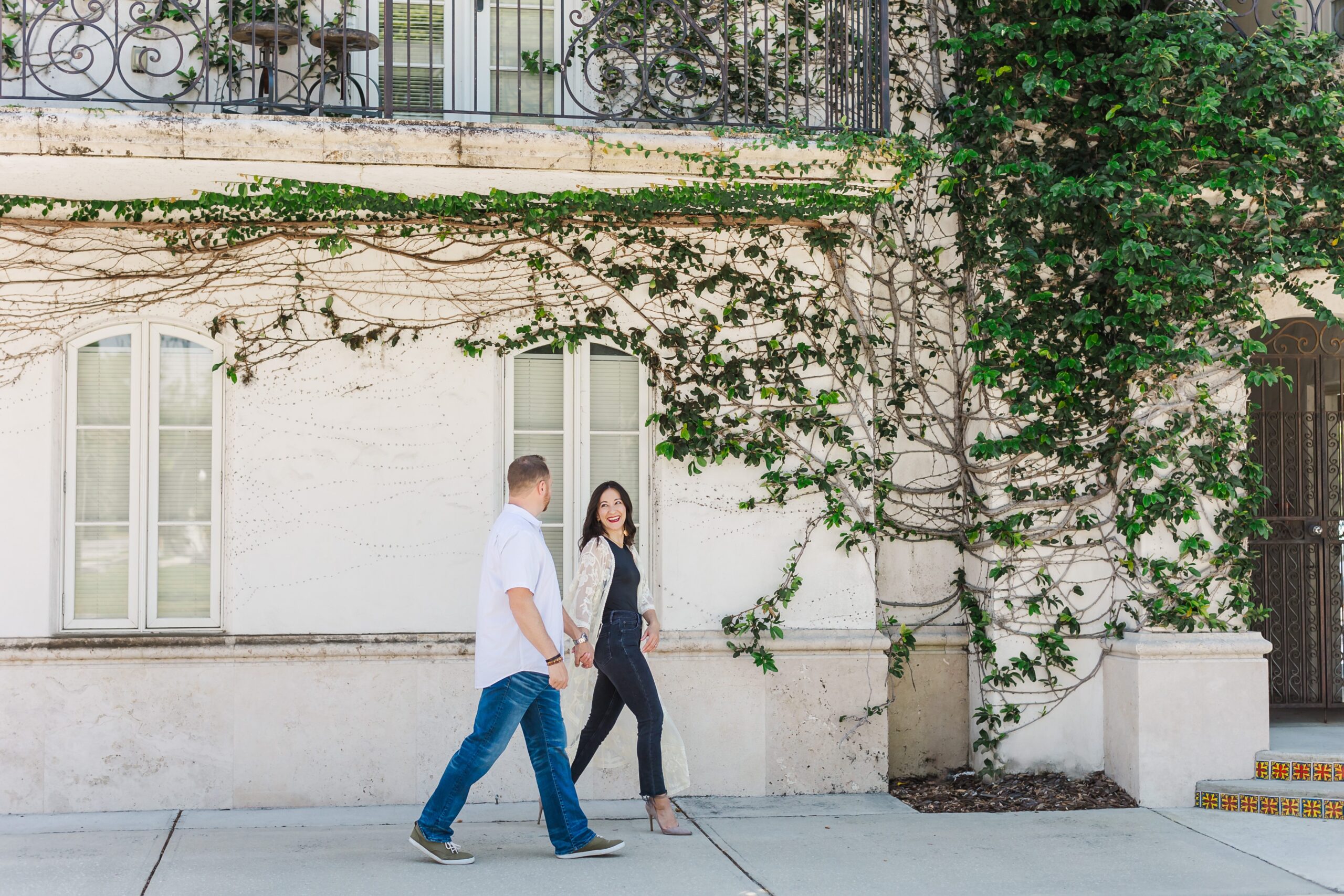 Couple walks holding hands in front of ivy wall at Hannibal Square in Winter Park, Florida for their engagement photos with Orlando Engagement Photographer Amy Britton Photography
