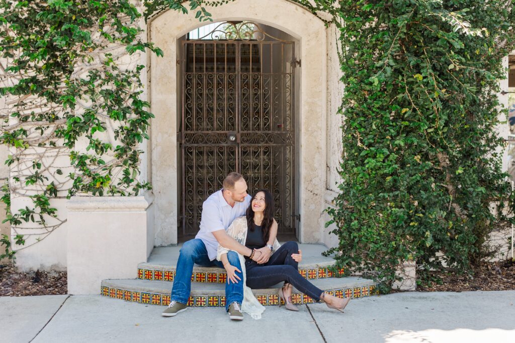 Couple sits on steps and smiles at each other in front of ivy wall at Hannibal Square in Winter Park, Florida for their engagement photos with Orlando Engagement Photographer Amy Britton Photography