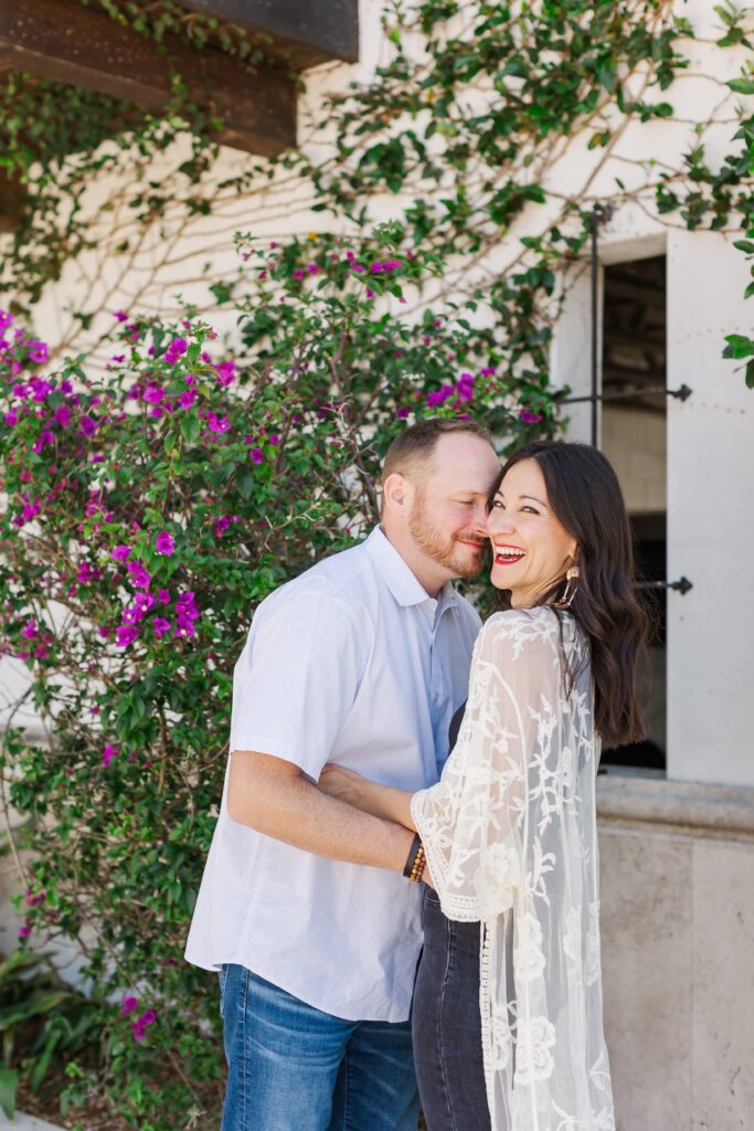 Guy snuggles into laughing girl in front of purple flowers and ivy wall at Hannibal Square in Winter Park, Florida for their engagement photos with Orlando Engagement Photographer Amy Britton Photography