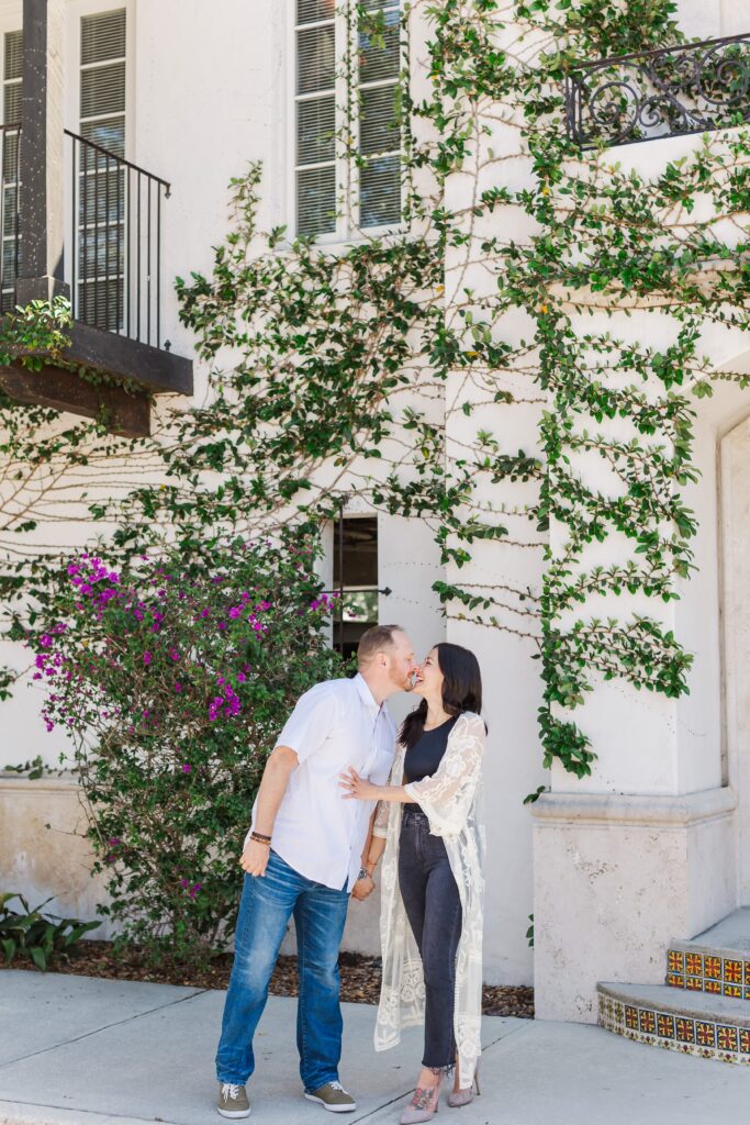 Couple kisses while holding hands in front of ivy wall with purple flowers at Hannibal Square in Winter Park, Florida for their engagement photos with Orlando Engagement Photographer Amy Britton Photography
