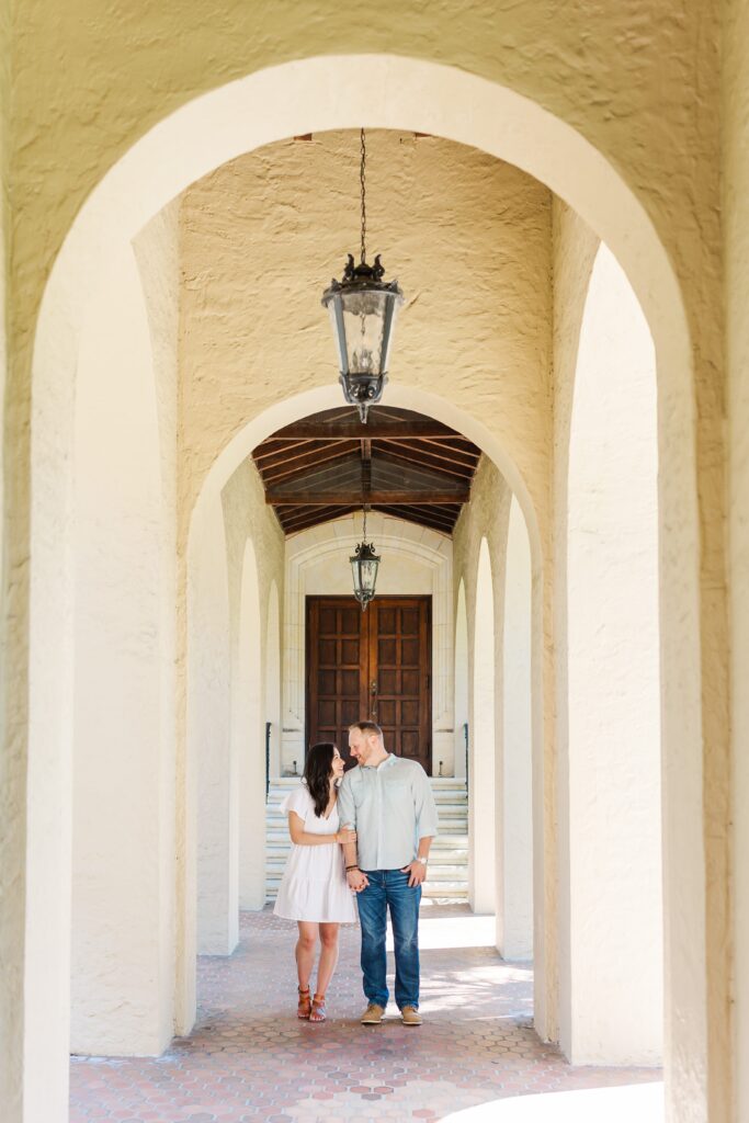 Couple wraps arms around each other in architectural hallway at Rollins College in Winter Park, Florida for their engagement photos with Orlando Engagement Photographer Amy Britton Photography