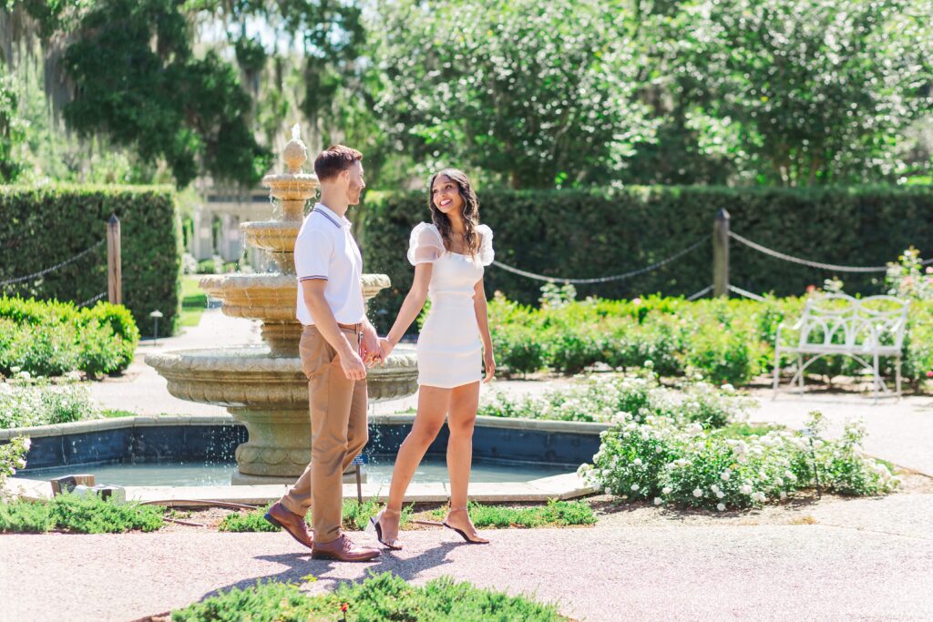 Couple holds hands and walks in front of fountain for their engagement photos in the rose garden at Leu Gardens in Orlando, Florida
