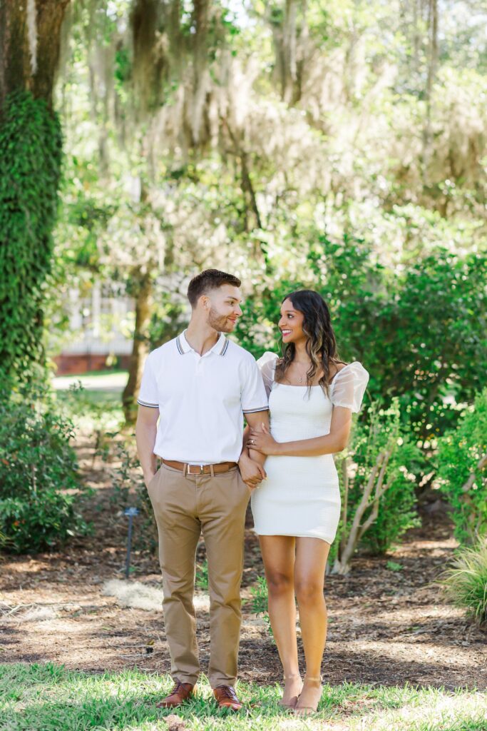 Couple looks at each other and smiles while girl holds guy's arm in front of large tree with hanging moss for their engagement photos at Leu Gardens in Orlando, Florida