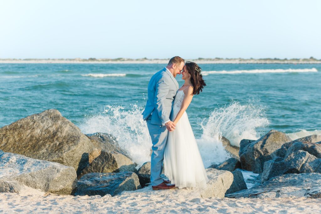 Bride and Groom hold hands as they stand on rocks from the jetty while waves splash at Vilano Beach for their St. Augustine beach wedding