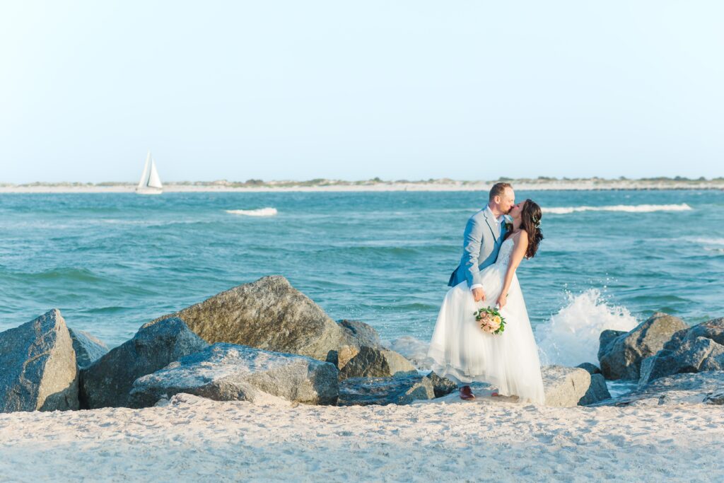 Groom dips and kisses Bride while she holds her bridal bouquet as they stand on rocks from the jetty at Vilano Beach with a sailboat in the distance for their St. Augustine beach wedding