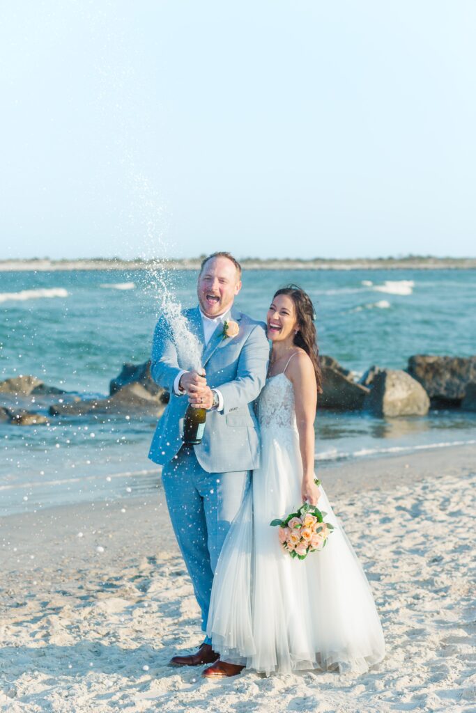 Bride and Groom pop champagne to celebrate their wedding as they stand on the beach with rocks from the jetty at Vilano Beach for their St. Augustine beach wedding