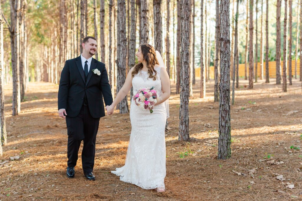 Bride and Groom hold hands and walk under pine trees for their forest wedding at The Pinery Orlando in Howey in the Hills, FL