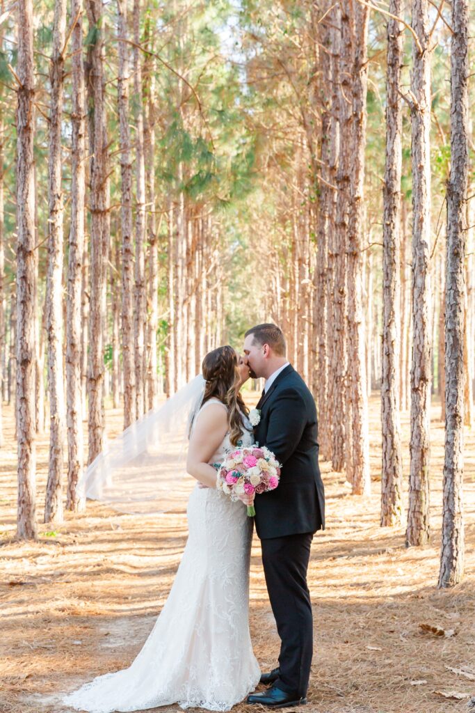 Bride and Groom kiss under pine trees while veil flies in the wind for their forest wedding at The Pinery Orlando in Howey in the Hills, FL
