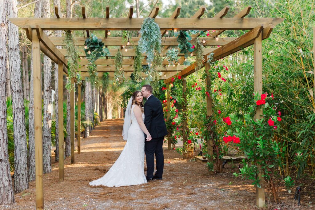 Groom kisses Bride's cheek under garden pergola with red flowers for their wedding at the Pinery Orlando Wedding and Events in Howey in the Hills, FL