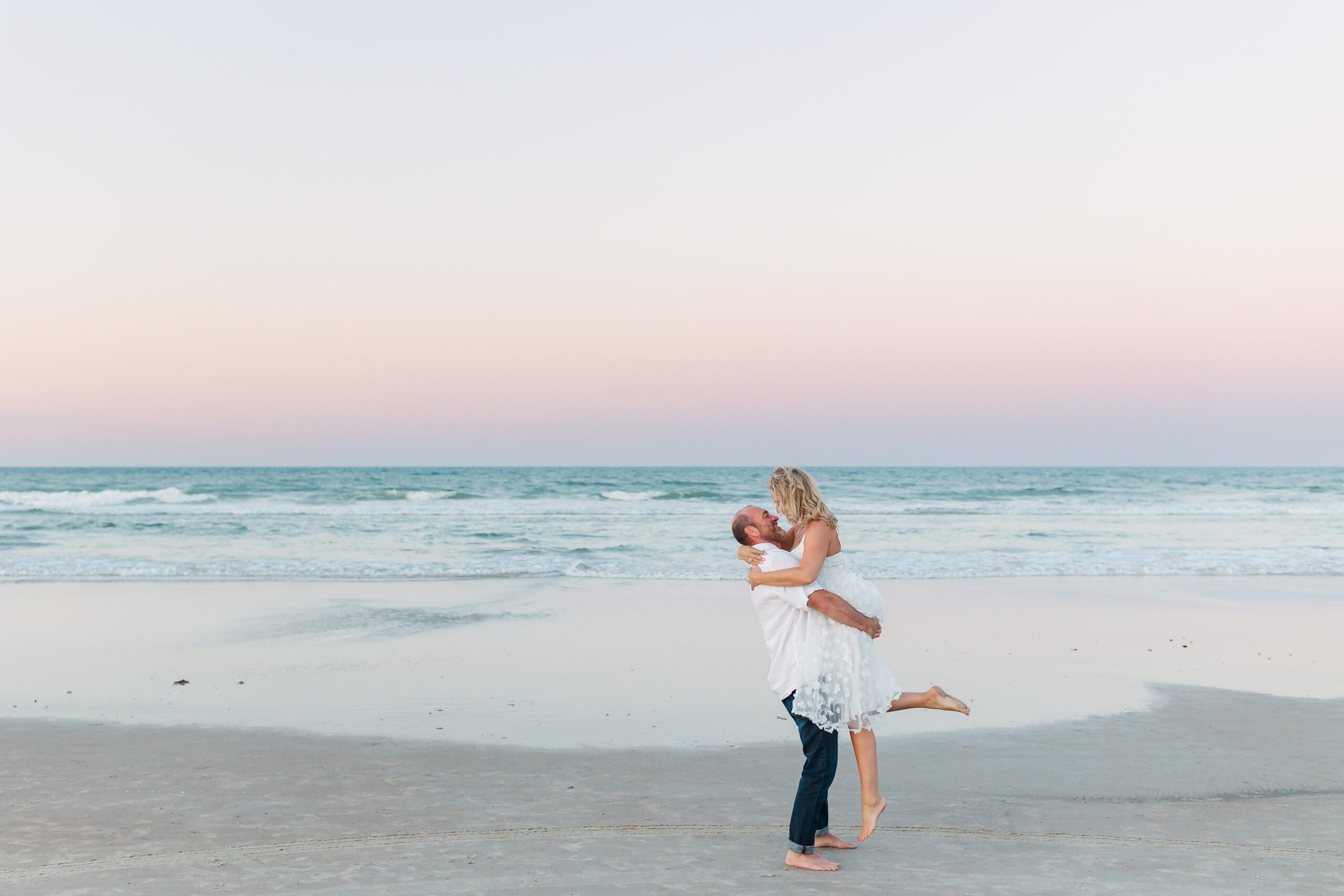 Guy lifts girl on the beach at sunset for their anniversary photos with their New Smyrna Beach Photographer