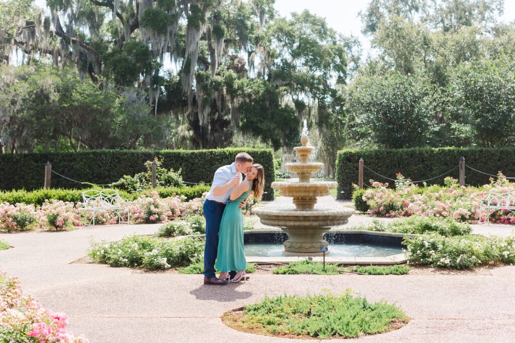 Guy kisses girl's cheek as they hold hands and stand in front of fountain in the rose garden for their engagement photos at Leu Gardens Orlando