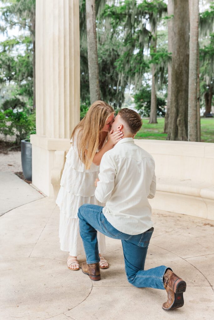 Girl kisses Guy as he proposes in front of the columns at Kraft Azalea Garden in Orlando, Florida