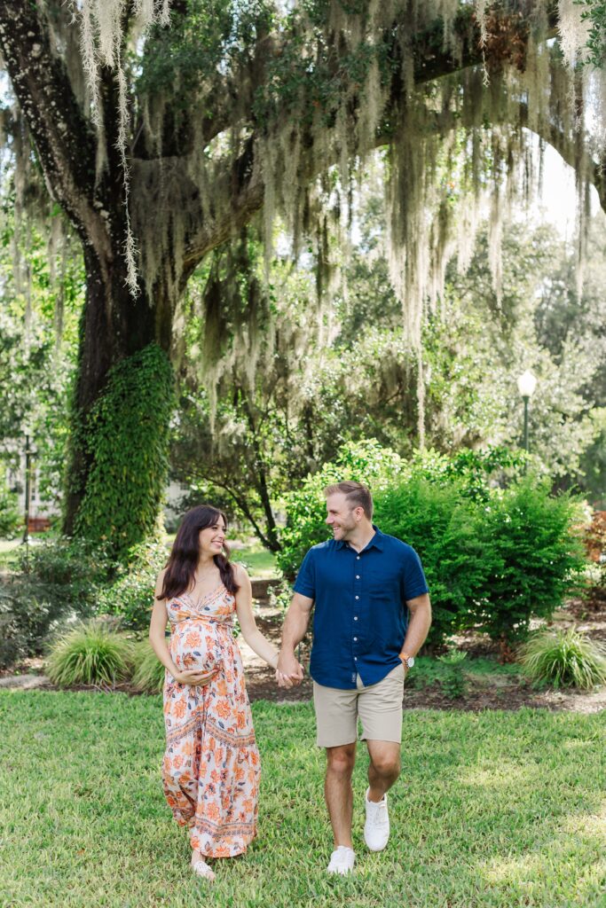 Couple walks under large tree with hanging moss for their Orlando Maternity Photos at Leu Gardens