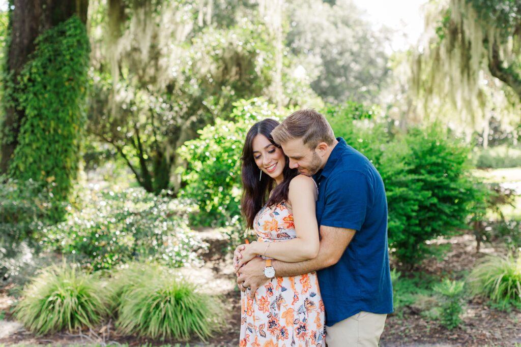 Guy wraps arms around girl under large tree with hanging moss for their Orlando Maternity Photos at Leu Gardens