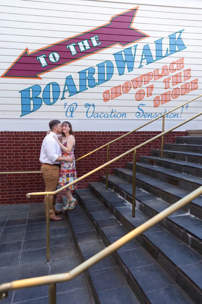 Guys kisses girl's cheek on the stairs in front of sign for their Disney Engagement Photos at The Boardwalk