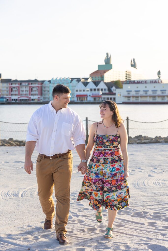 Couple holds hands and walks along the beach at Disney's Beach Club Resort for their engagement photos