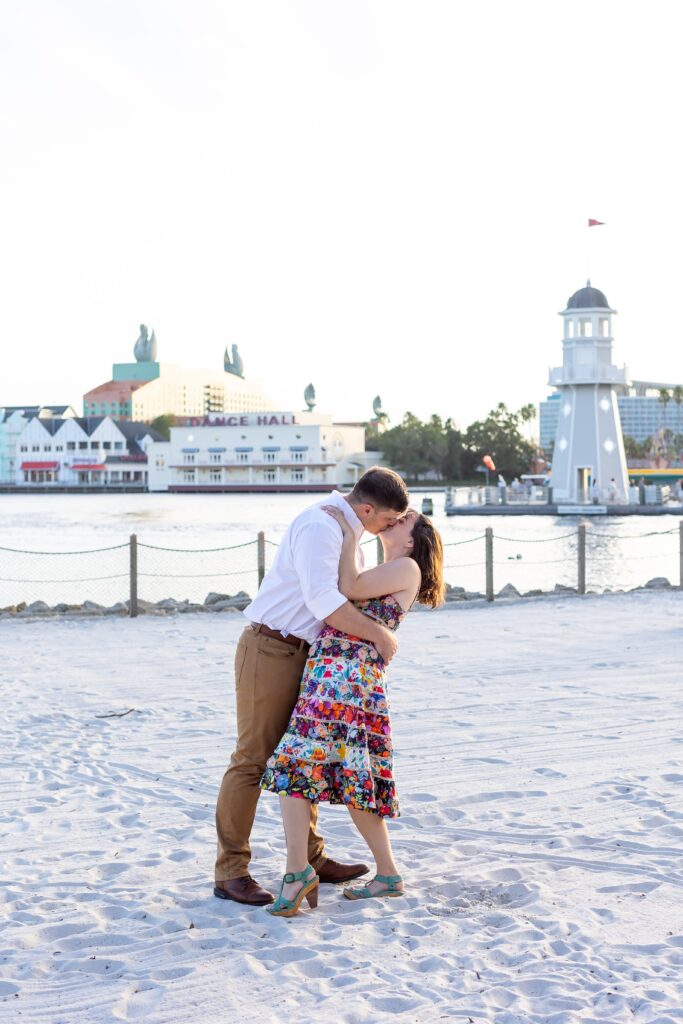 Guy dips and kisses girl in front of lighthouse on the beach at Disney's Beach Club Resort for their engagement photos