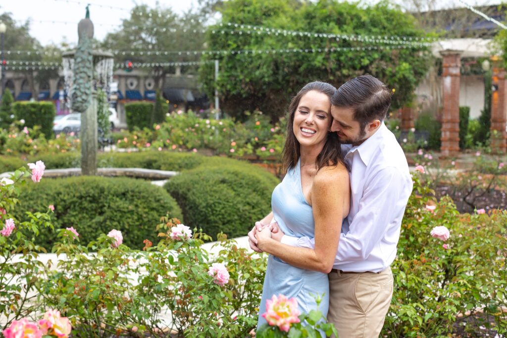 Couple smiles and snuggles in roses in front of peacock fountain for their engagement photos on Park Ave in Winter Park, Florida