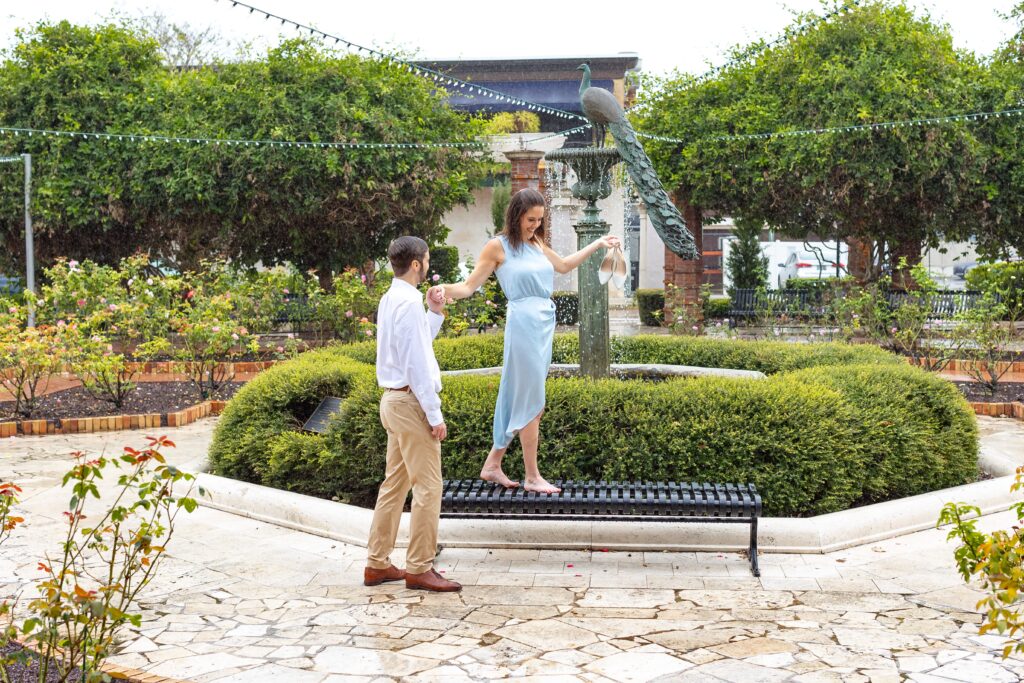 Girl walks on bench as guy holds her hand front of peacock fountain for their engagement photos on Park Ave in Winter Park, Florida