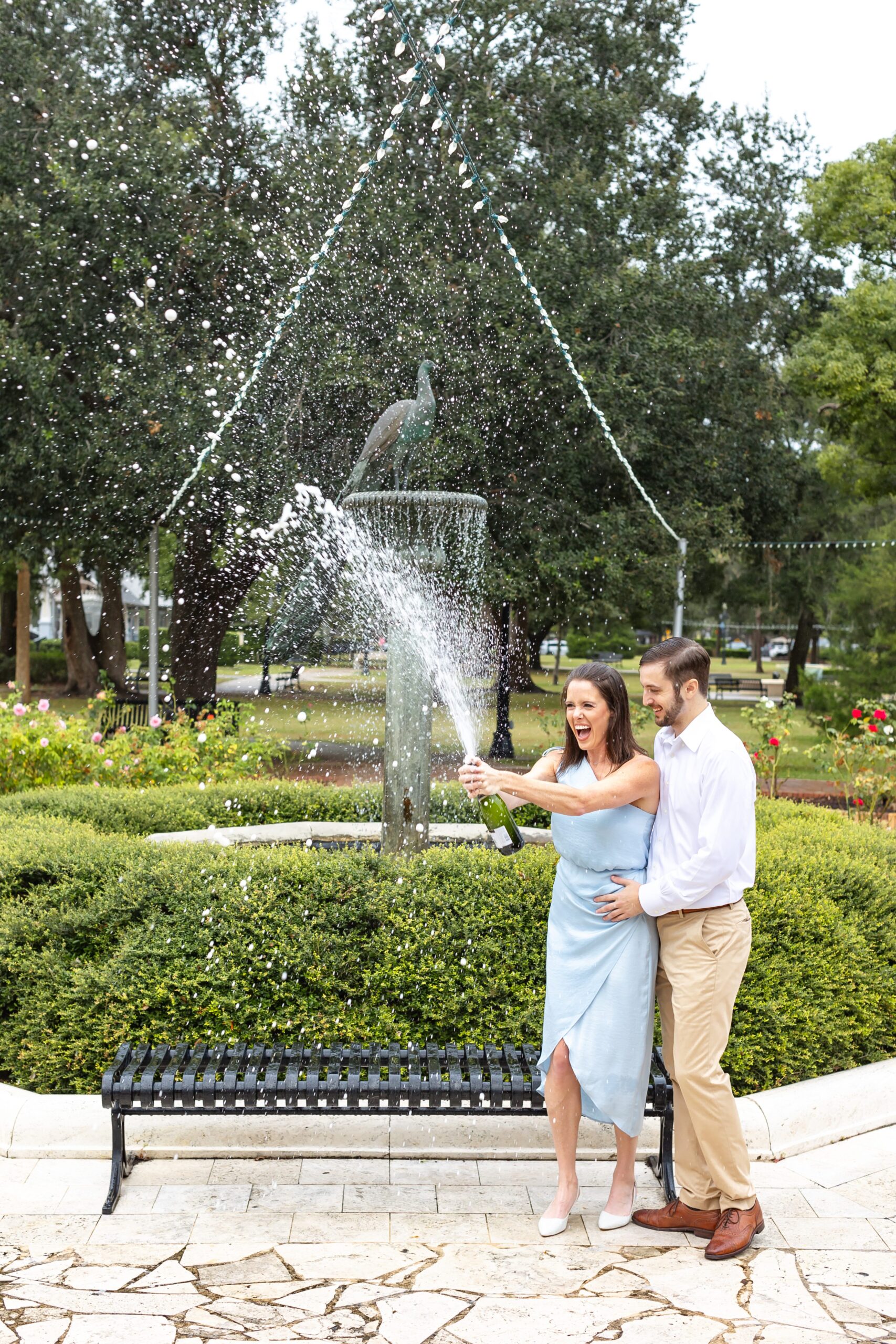 Couple pops champagne in front of peacock fountain for their engagement photos on Park Ave in Winter Park, Florida