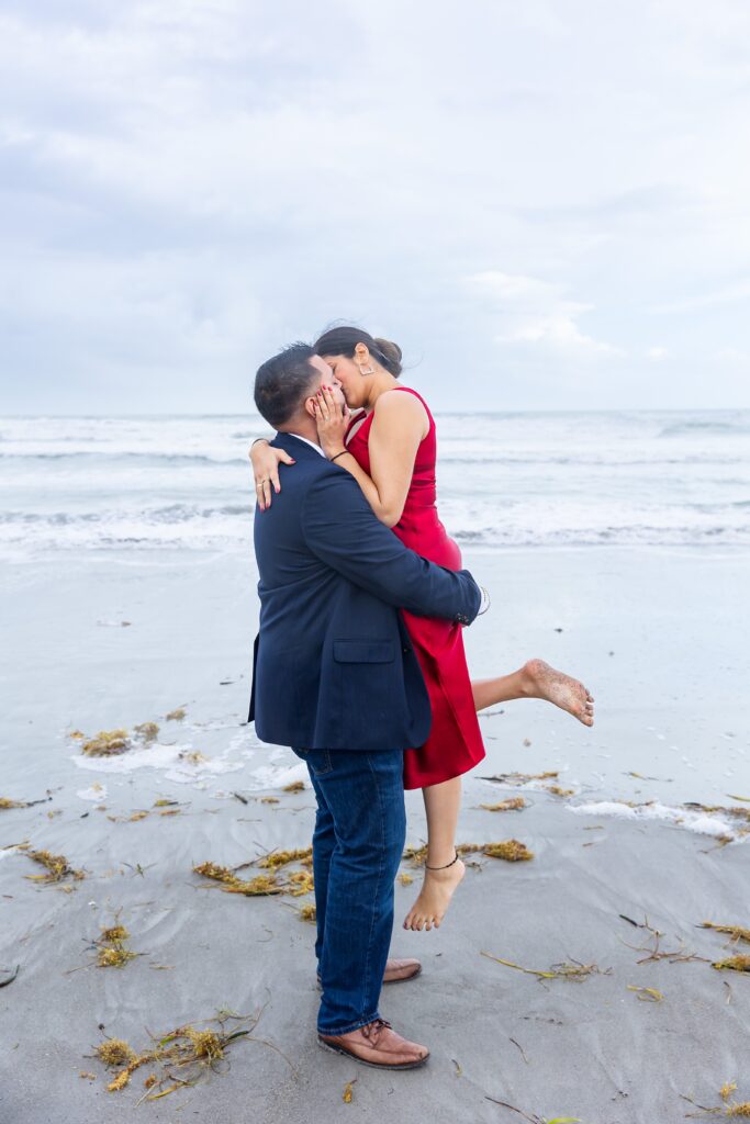 Guy lifts girl on and kisses her in front of the ocean at Lori Wilson Park for their engagement photos in Cocoa Beach, Florida