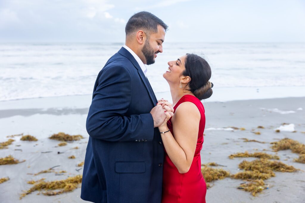 Couple holds hands and laughs in front of the ocean at Lori Wilson Park for their engagement photos in Cocoa Beach, Florida