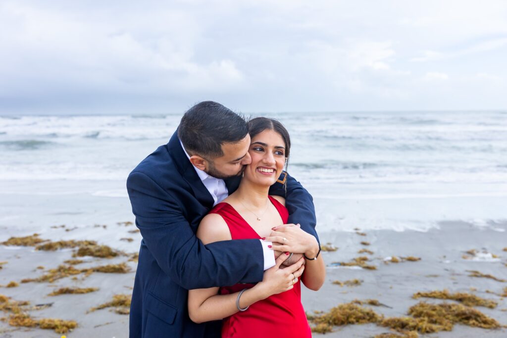 Guy wraps arms around girl on and kisses her in front of the ocean at Lori Wilson Park for their engagement photos in Cocoa Beach, Florida