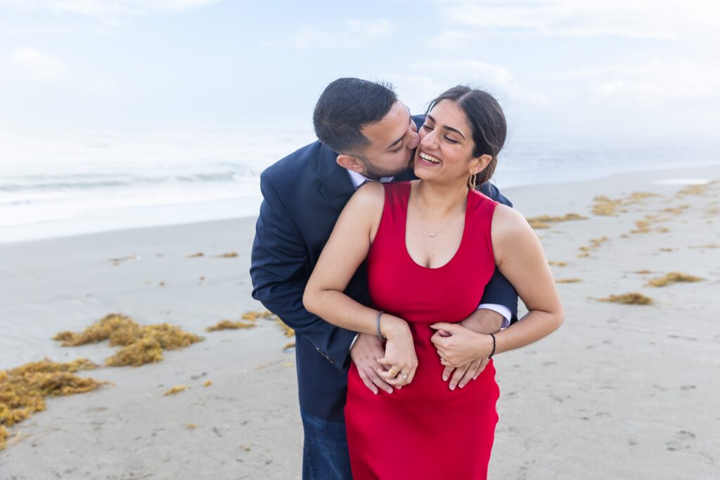 Guy wraps arms around girl on and kisses her in front of the ocean at Lori Wilson Park for their engagement photos in Cocoa Beach, Florida