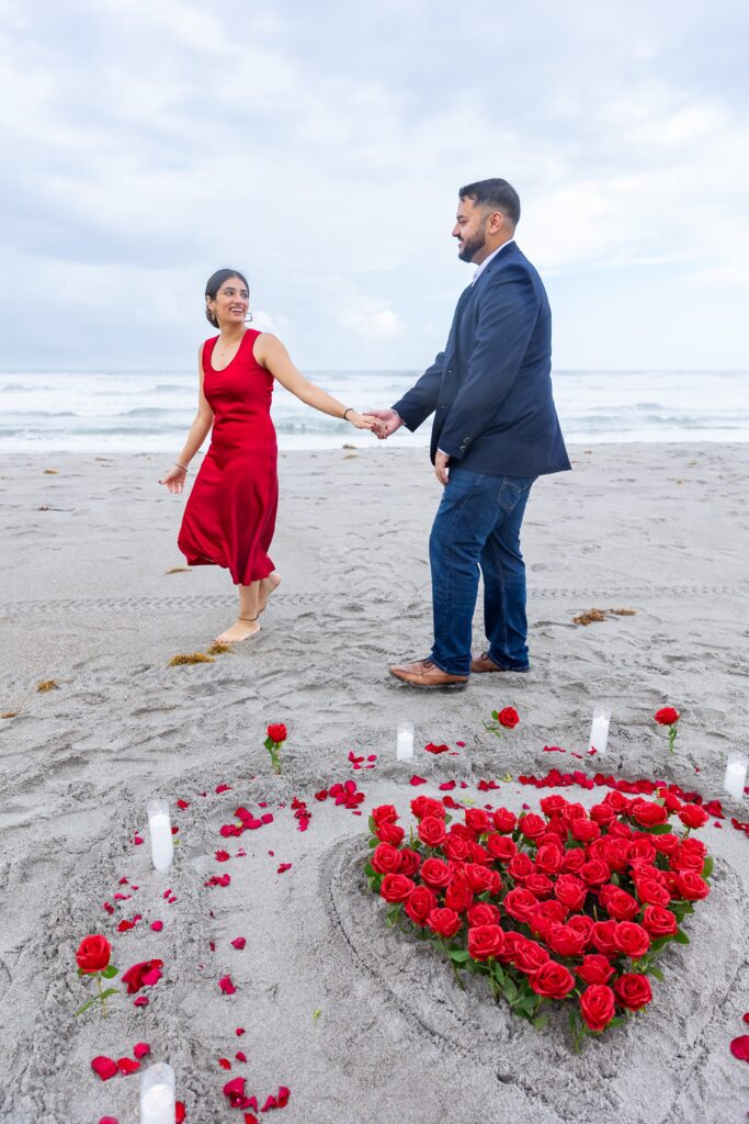 Couple holds hands and walks in front of heart shaped roses in front of the ocean at Lori Wilson Park for their engagement photos in Cocoa Beach, Florida