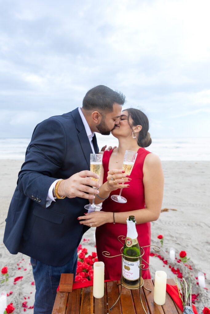 Couple kisses with champagne after their proposal at Lori Wilson Park for their engagement photos in Cocoa Beach, Florida