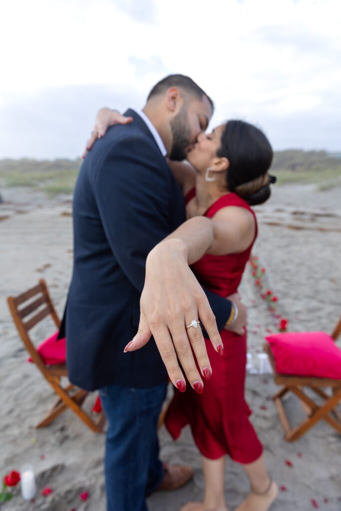 Couple kisses and girl holds out ring after their proposal at Lori Wilson Park for their engagement photos in Cocoa Beach, Florida