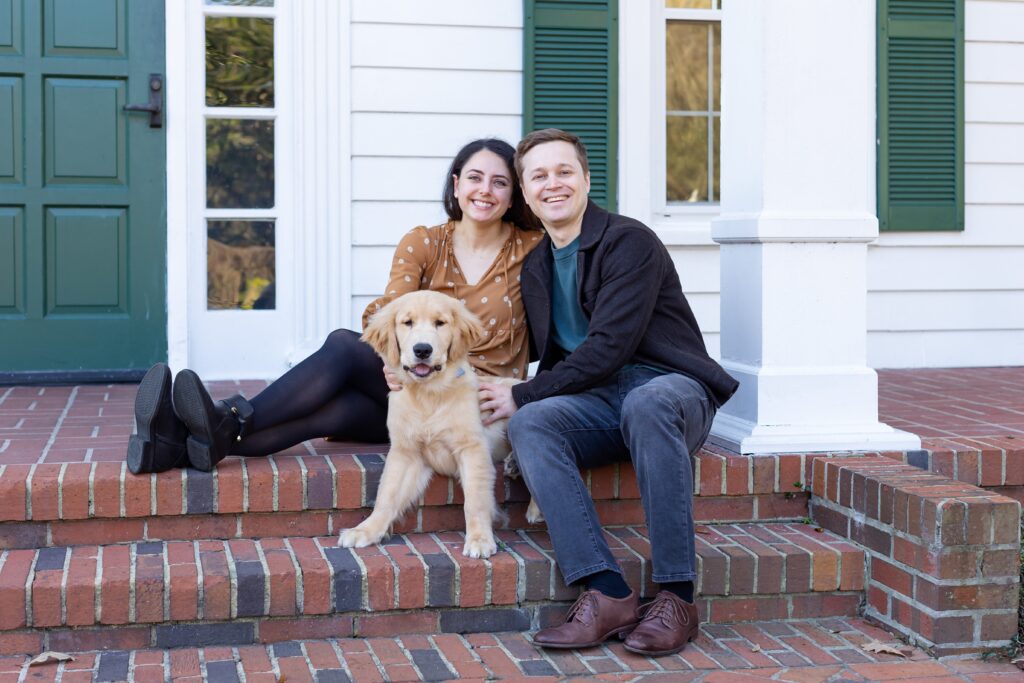 Couple poses with their dog in front of Cypress Grove Estate House for their engagement photos at Cypress Grove Park in Orlando, Florida