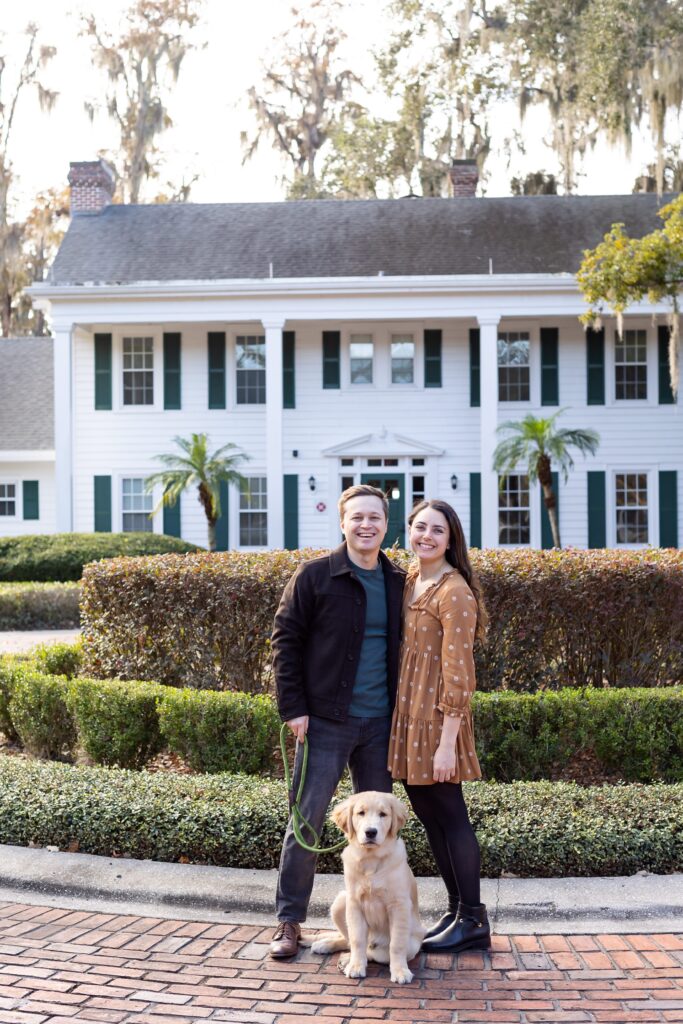 Couple poses with their dog in front of Cypress Grove Estate House for their engagement photos at Cypress Grove Park in Orlando, Florida