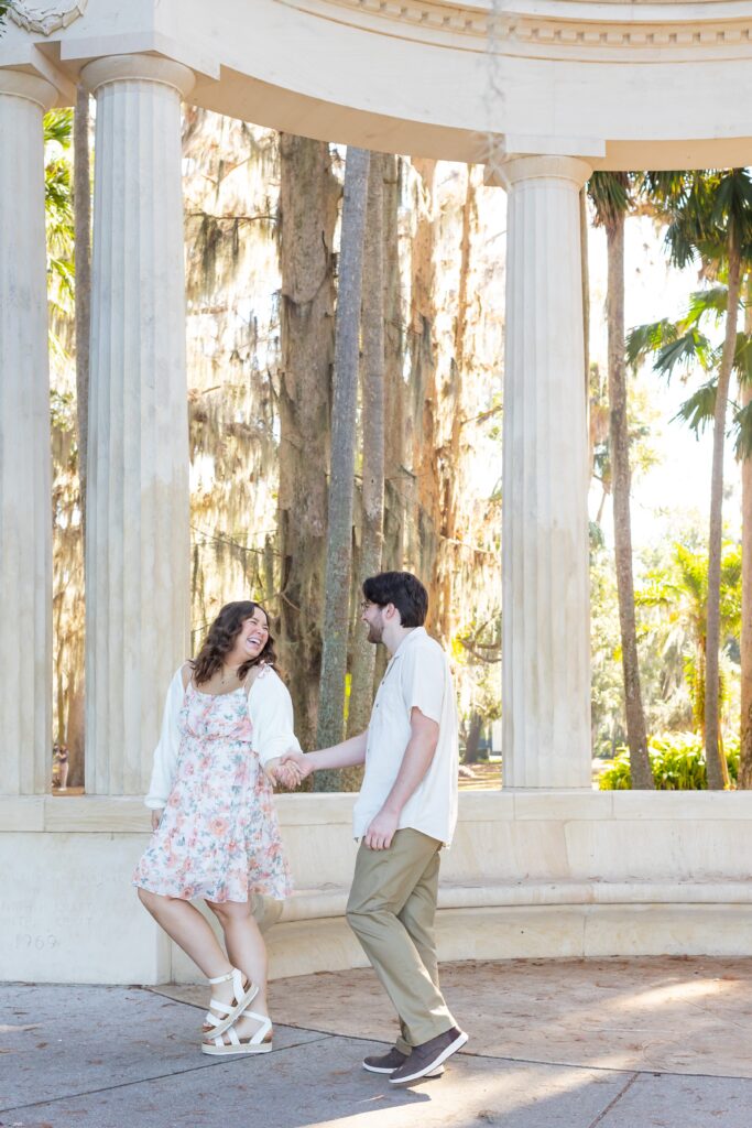 Couple holds hands and walks under the columns for their engagement photos at Kraft Azalea Garden in Winter Park, Florida