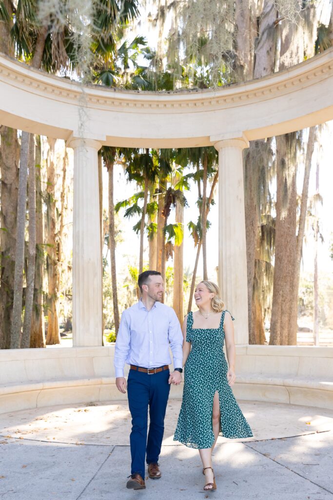 Couple holds hands and walks in front of columns for their engagement photos at Kraft Azalea Garden in Orlando, Florida