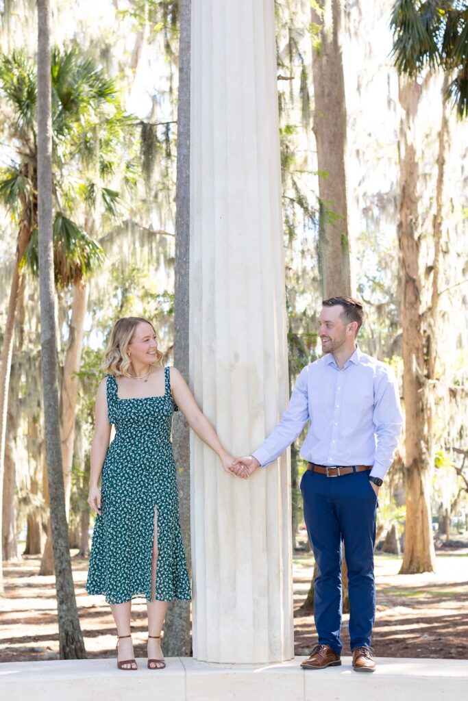 Couple holds hands and smiles at each other on top of columns for their engagement photos at Kraft Azalea Garden in Orlando, Florida