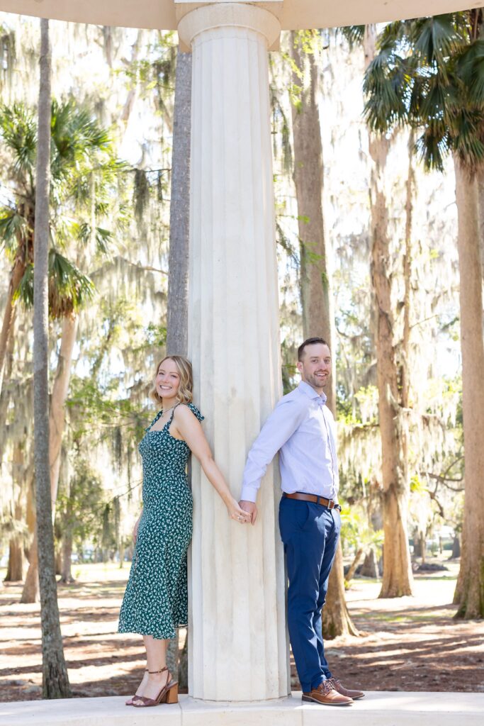 Couple holds hands and smiles on top of columns for their engagement photos at Kraft Azalea Garden in Orlando, Florida